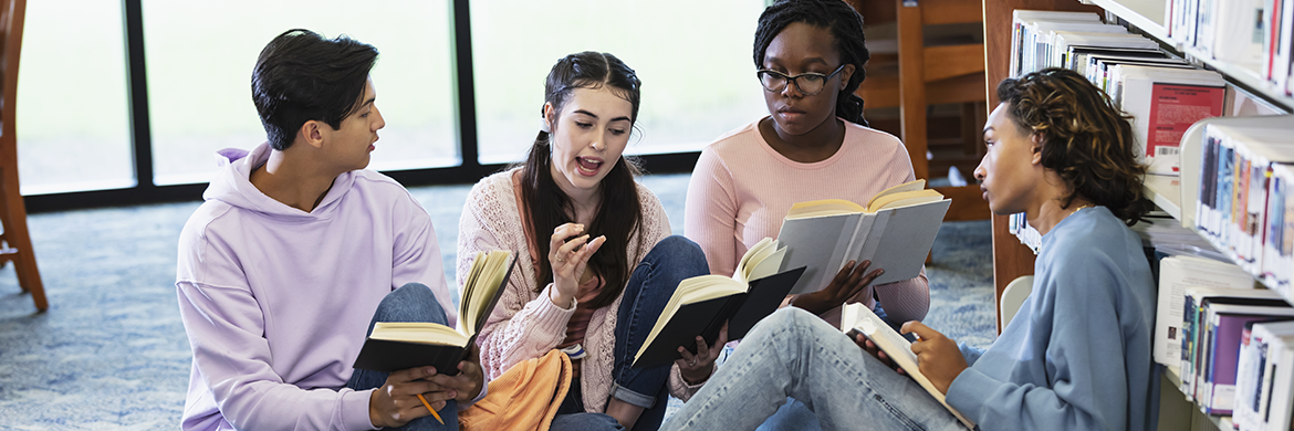 Group of teens sitting in the library with open books during what looks to be a discussion